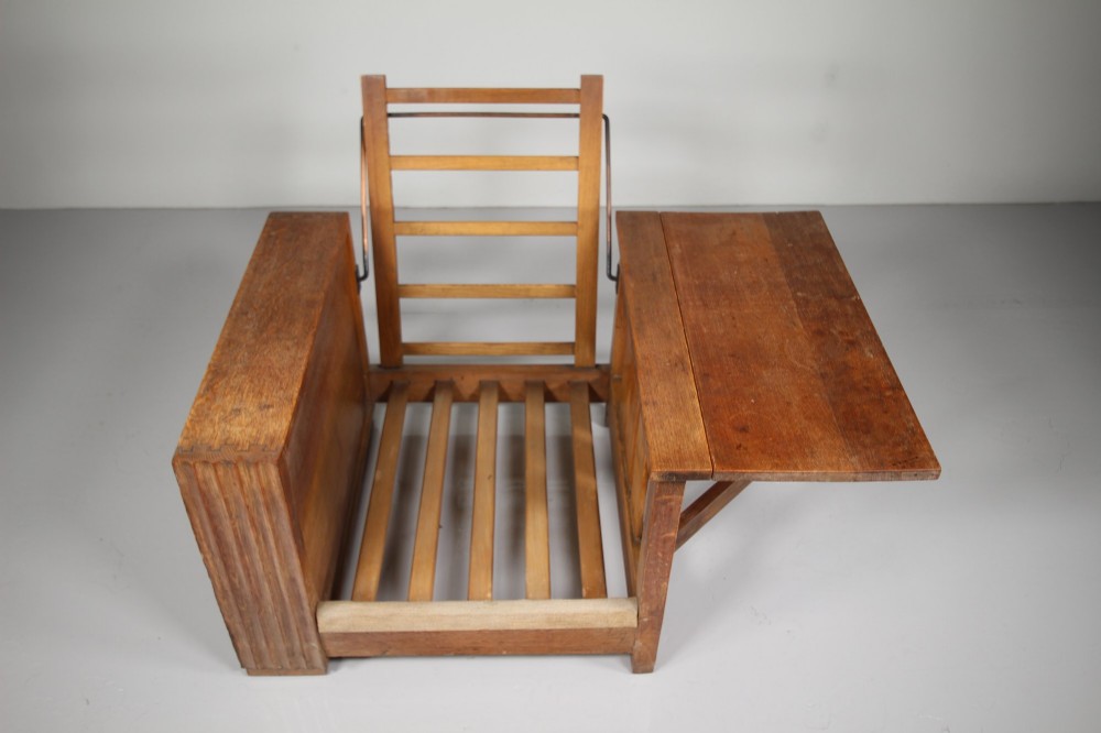 A 1930s solid oak reclining armchair frame featuring integrated side bookshelves, an adjustable slatted backrest, and a folding drop-leaf side table. View IX