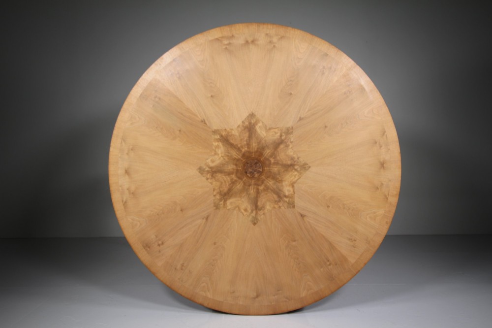 An English Art Deco circular dining table crafted from light timber with a veneered top supported by a curved four-leg pedestal base and cross stretcher. View VI