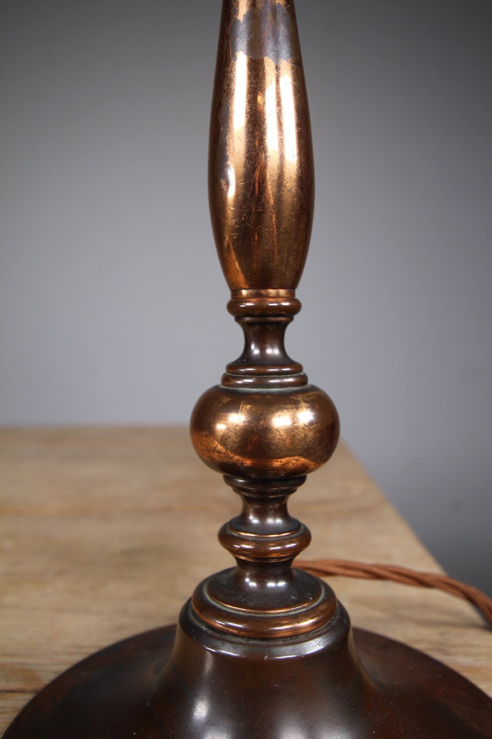 An antique coppered metal table lamp base featuring a turned baluster stem, a stepped circular foot, and a brass bulb fitting. View II