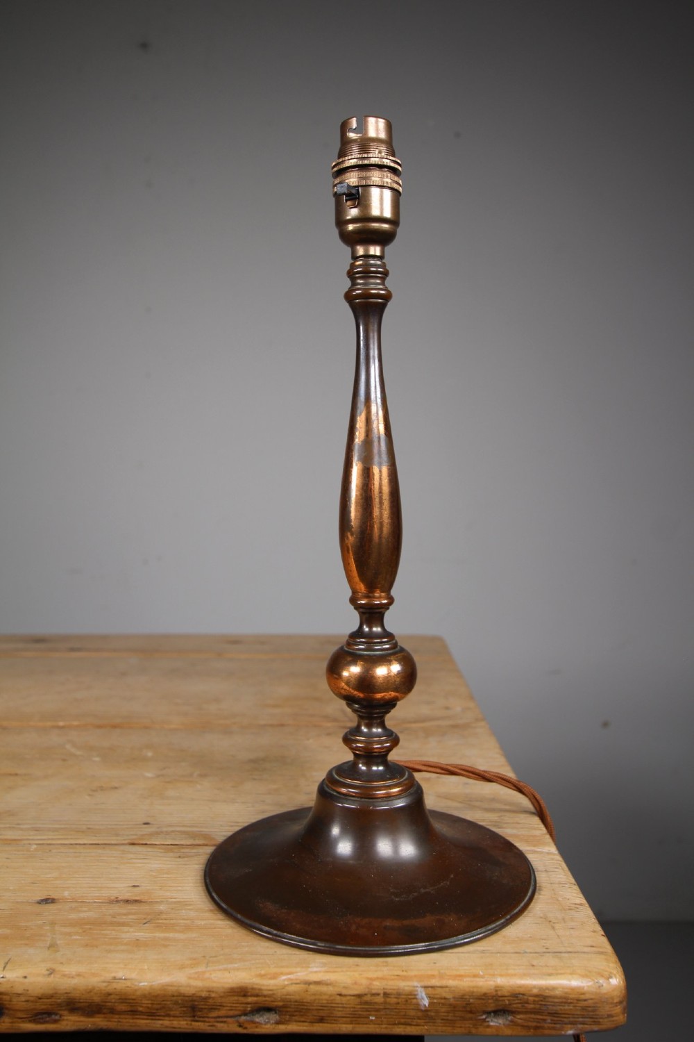 An antique coppered metal table lamp base featuring a turned baluster stem, a stepped circular foot, and a brass bulb fitting. View V