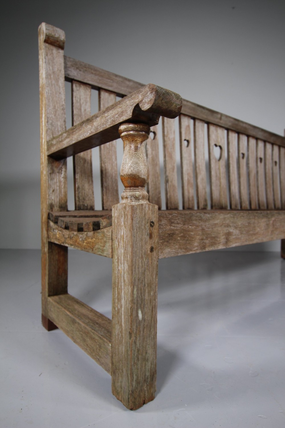 An antique weathered teak garden bench featuring a slatted backrest with pierced heart motifs and turned armrest supports. View II
