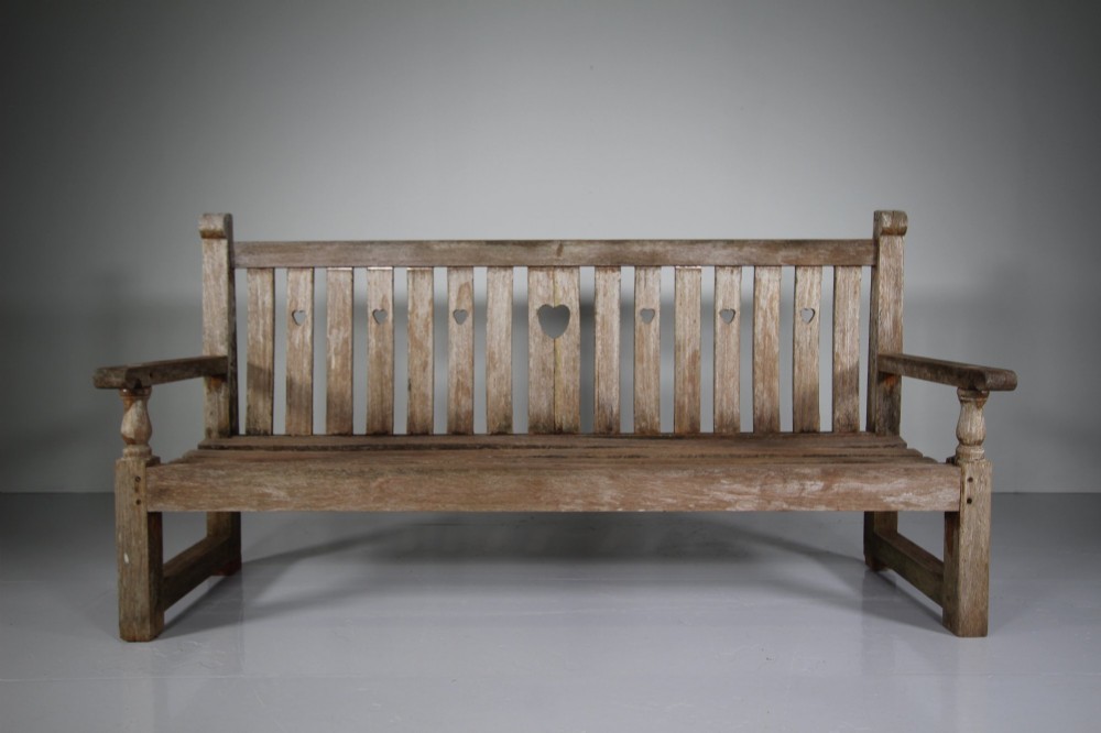 An antique weathered teak garden bench featuring a slatted backrest with pierced heart motifs and turned armrest supports.