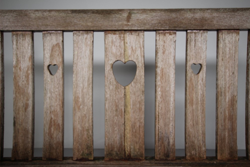 An antique weathered teak garden bench featuring a slatted backrest with pierced heart motifs and turned armrest supports. View III