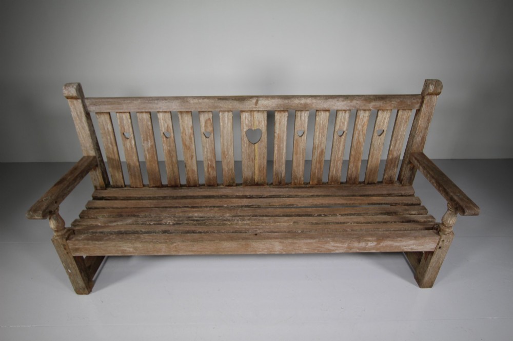 An antique weathered teak garden bench featuring a slatted backrest with pierced heart motifs and turned armrest supports. View IV