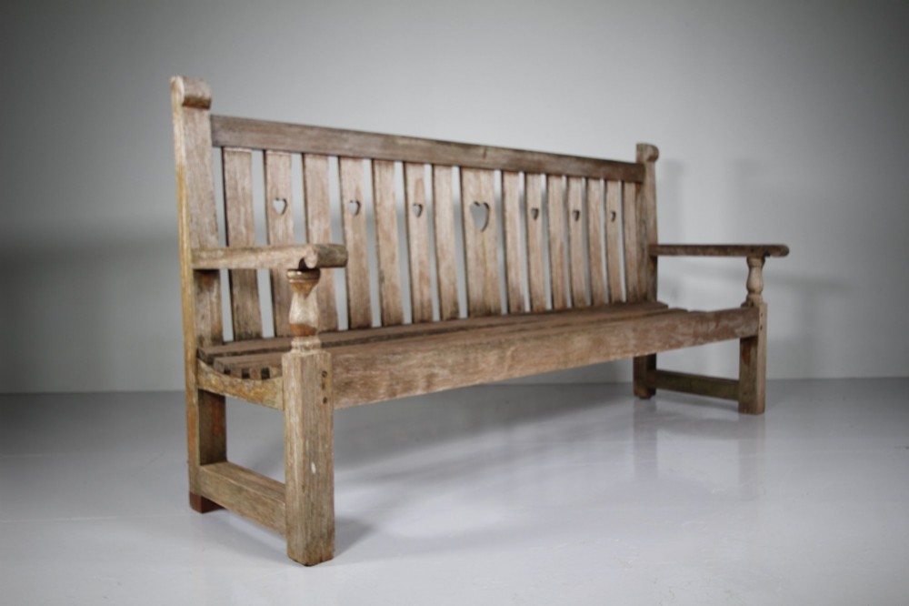 An antique weathered teak garden bench featuring a slatted backrest with pierced heart motifs and turned armrest supports. View V
