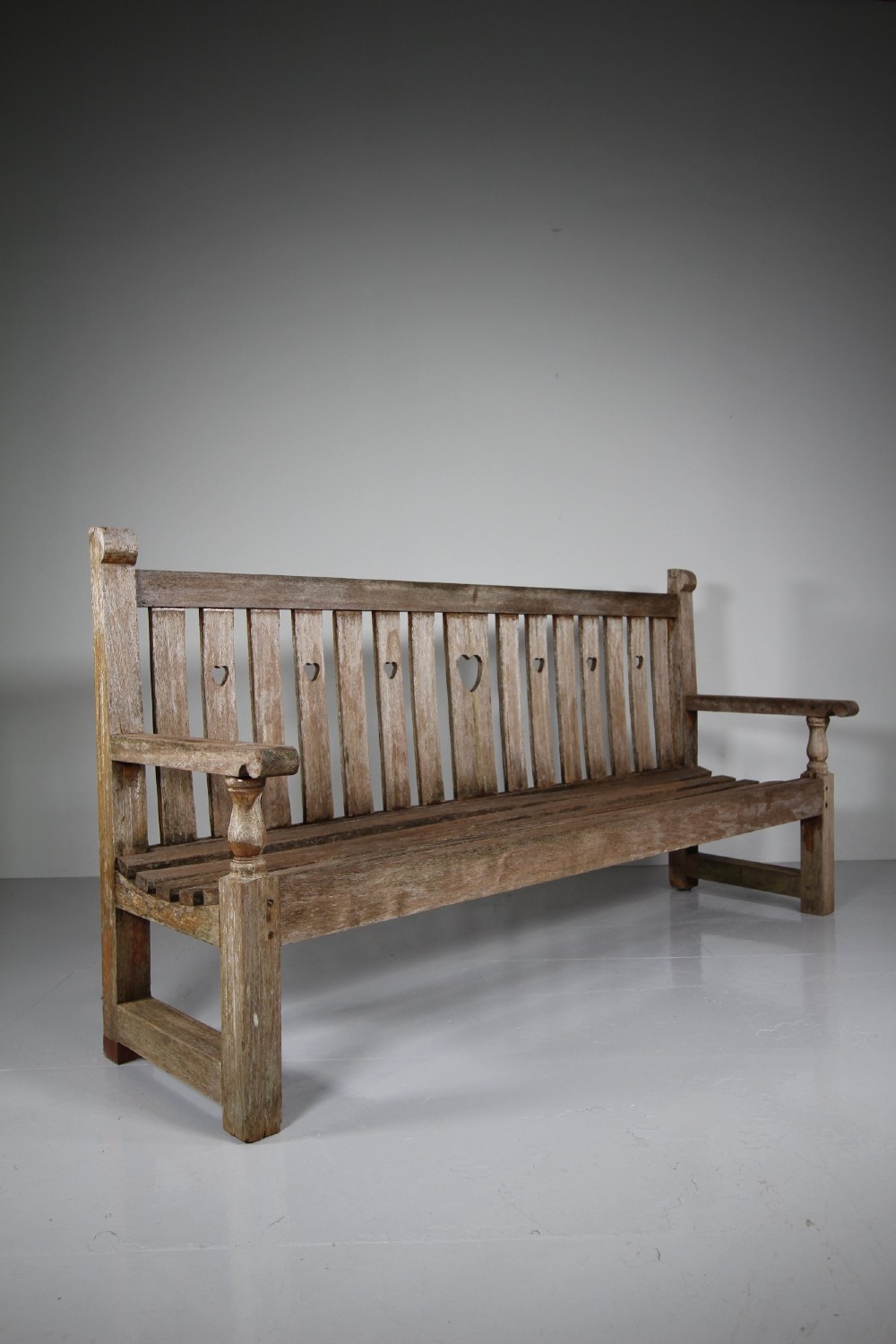 An antique weathered teak garden bench featuring a slatted backrest with pierced heart motifs and turned armrest supports. View VI