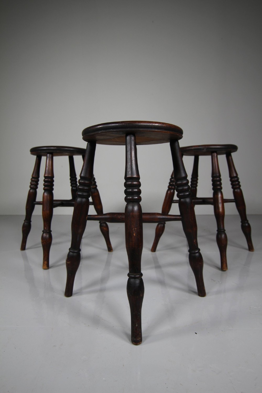 A set of three 19th-century dark brown wooden kitchen stools featuring circular seats and turned splayed legs with stretchers. View V