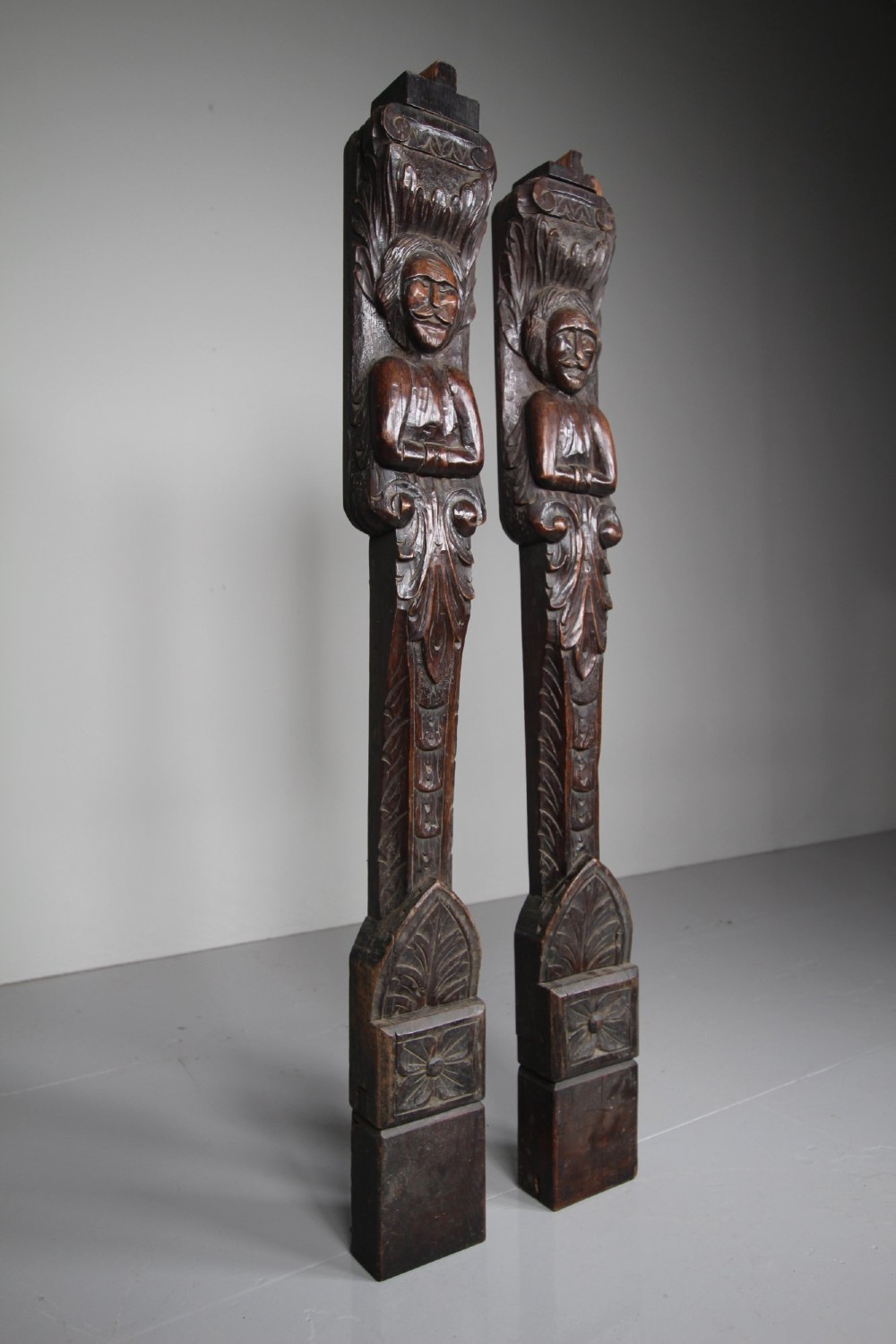 A pair of vertical hand-carved dark oak pilasters featuring stylized human figures at the top above foliage and geometric carved motifs.