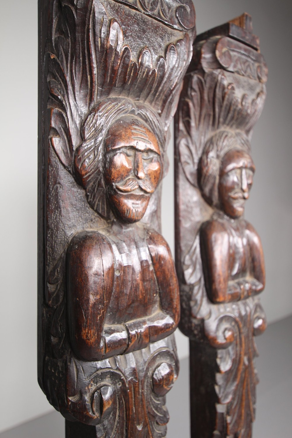 A pair of vertical hand-carved dark oak pilasters featuring stylized human figures at the top above foliage and geometric carved motifs. View II