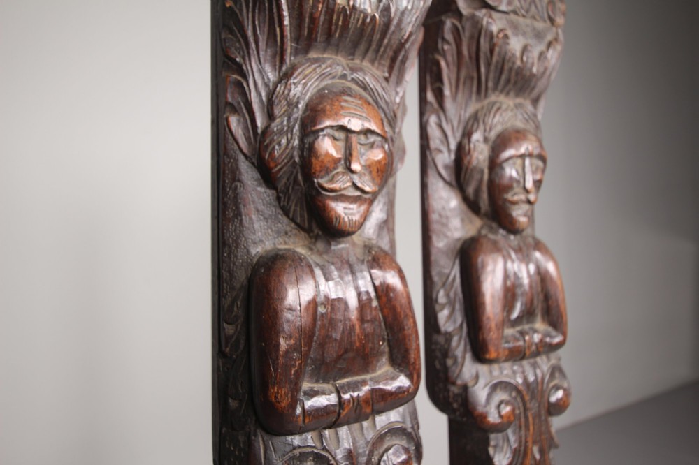 A pair of vertical hand-carved dark oak pilasters featuring stylized human figures at the top above foliage and geometric carved motifs. View III