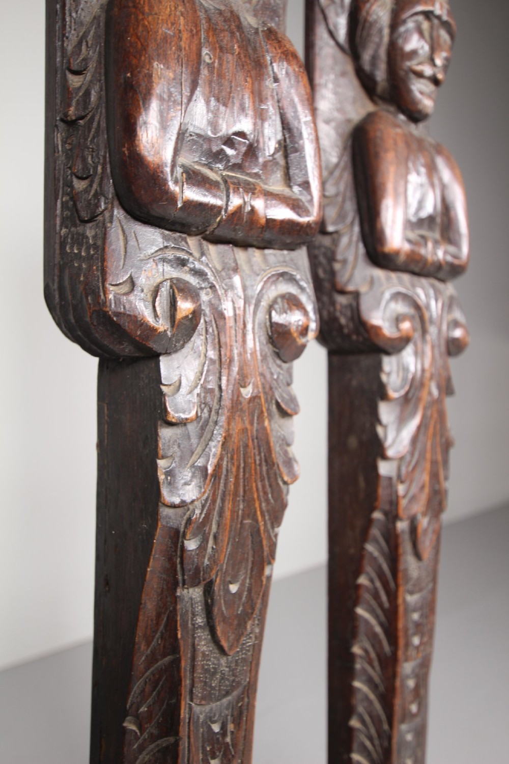 A pair of vertical hand-carved dark oak pilasters featuring stylized human figures at the top above foliage and geometric carved motifs. View V