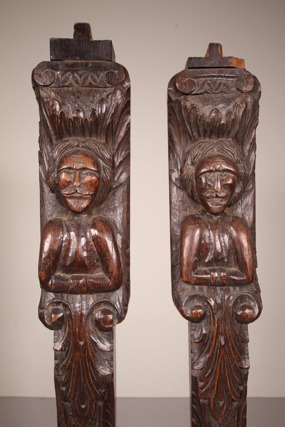 A pair of vertical hand-carved dark oak pilasters featuring stylized human figures at the top above foliage and geometric carved motifs. View VII