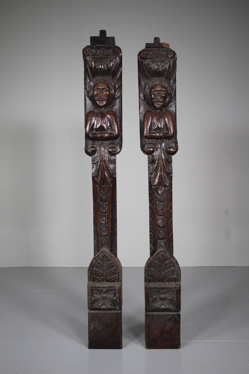 A pair of vertical hand-carved dark oak pilasters featuring stylized human figures at the top above foliage and geometric carved motifs. View VIII