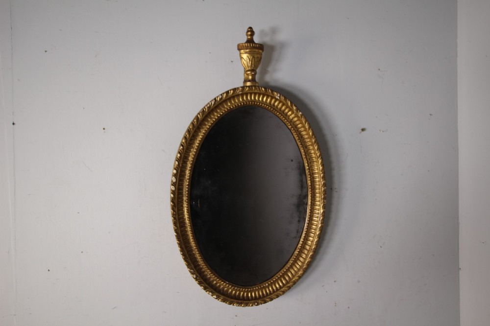 An 18th-century Neoclassical oval giltwood wall mirror featuring an urn finial top and fluted border along the carved frame.