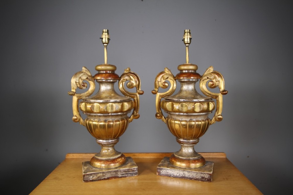 A pair of 19th-century carved pine urn table lamps with distressed gold leaf finish, scrolled handles, and square plinth bases.