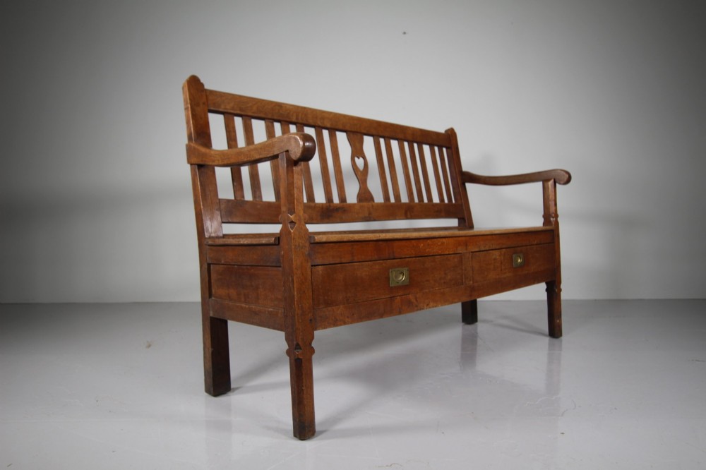A nineteenth-century oak hall bench with a slatted back featuring a central pierced heart motif, scrolled arms, and two drawers with flush brass handles beneath the seat. View V