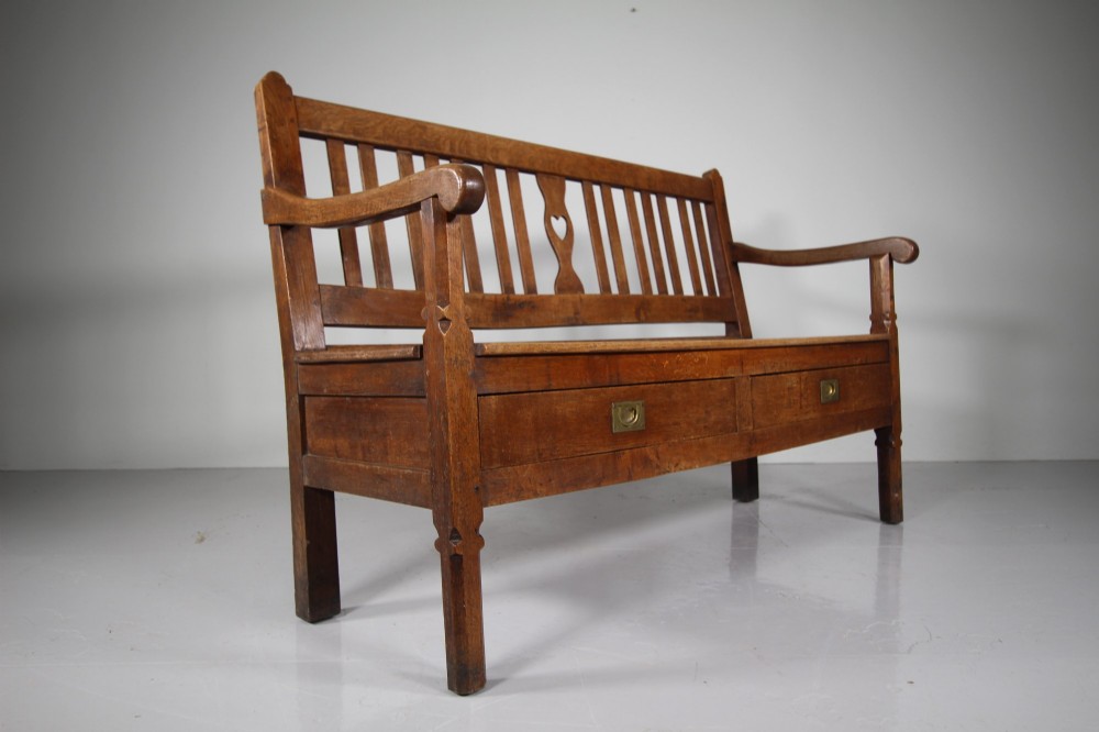 A nineteenth-century oak hall bench with a slatted back featuring a central pierced heart motif, scrolled arms, and two drawers with flush brass handles beneath the seat. View VI