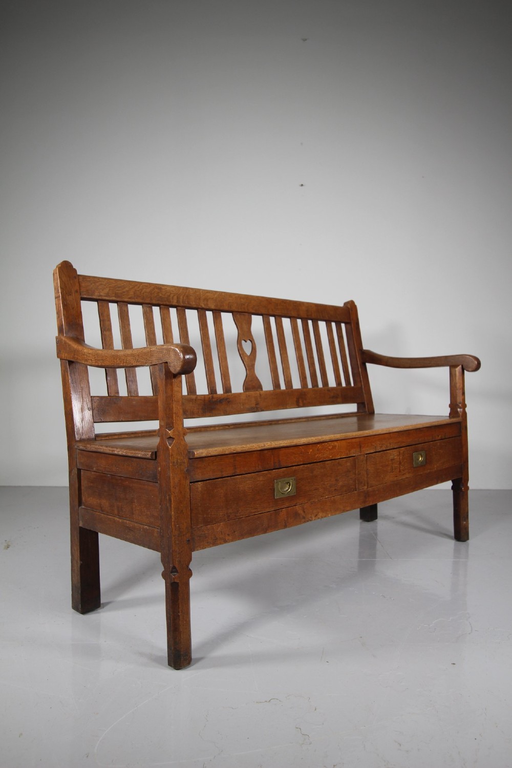 A nineteenth-century oak hall bench with a slatted back featuring a central pierced heart motif, scrolled arms, and two drawers with flush brass handles beneath the seat. View VII