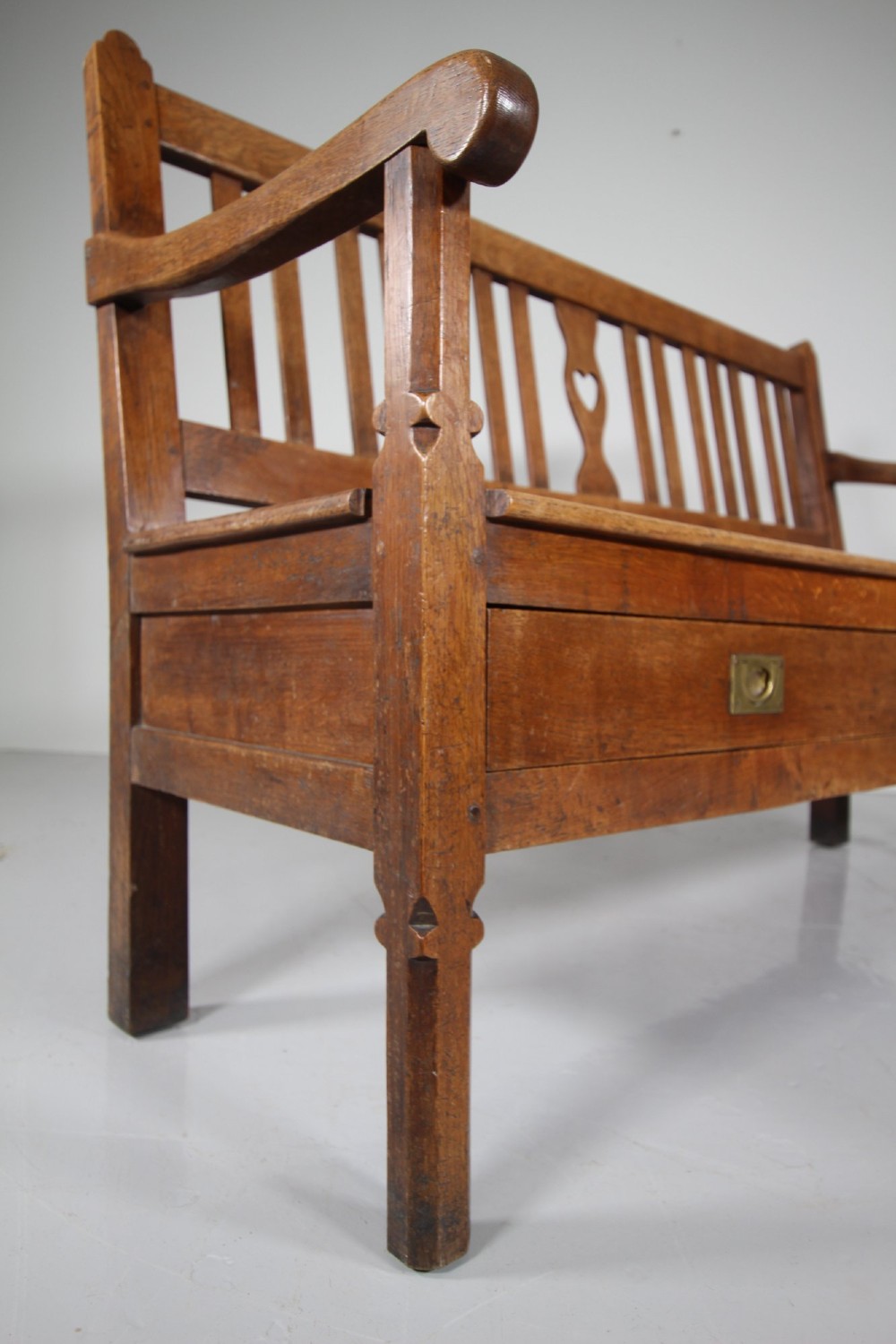 A nineteenth-century oak hall bench with a slatted back featuring a central pierced heart motif, scrolled arms, and two drawers with flush brass handles beneath the seat. View VIII