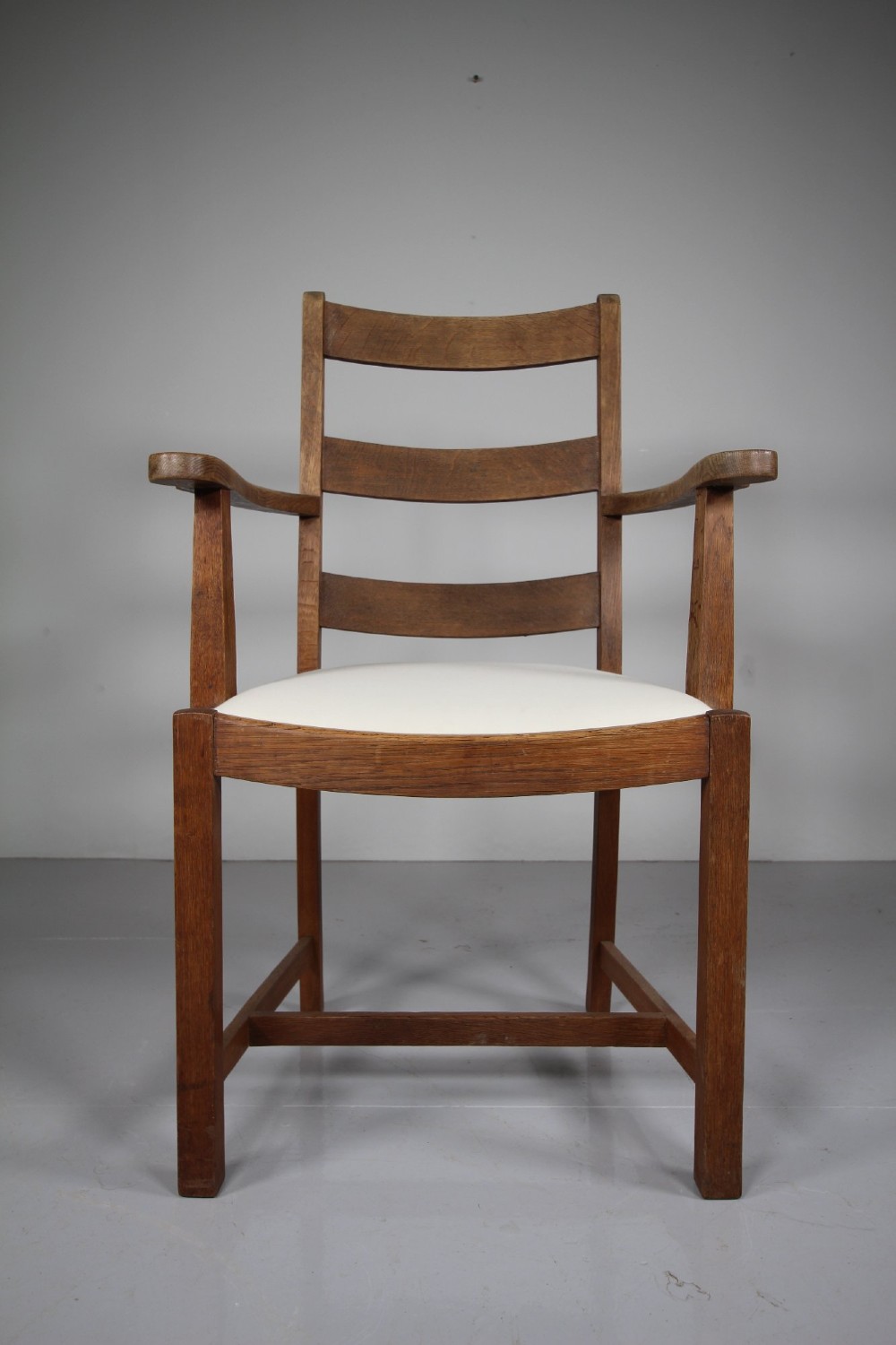 An Arts and Crafts solid oak armchair featuring a ladderback design, flat armrests, and an upholstered cream seat cushion. View V