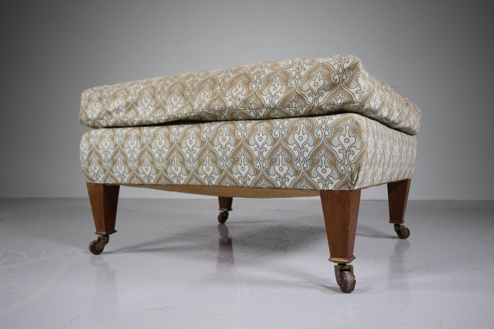 A large square antique footstool upholstered in a beige patterned fabric with a loose top cushion, supported by four tapered wooden legs on brass castors.