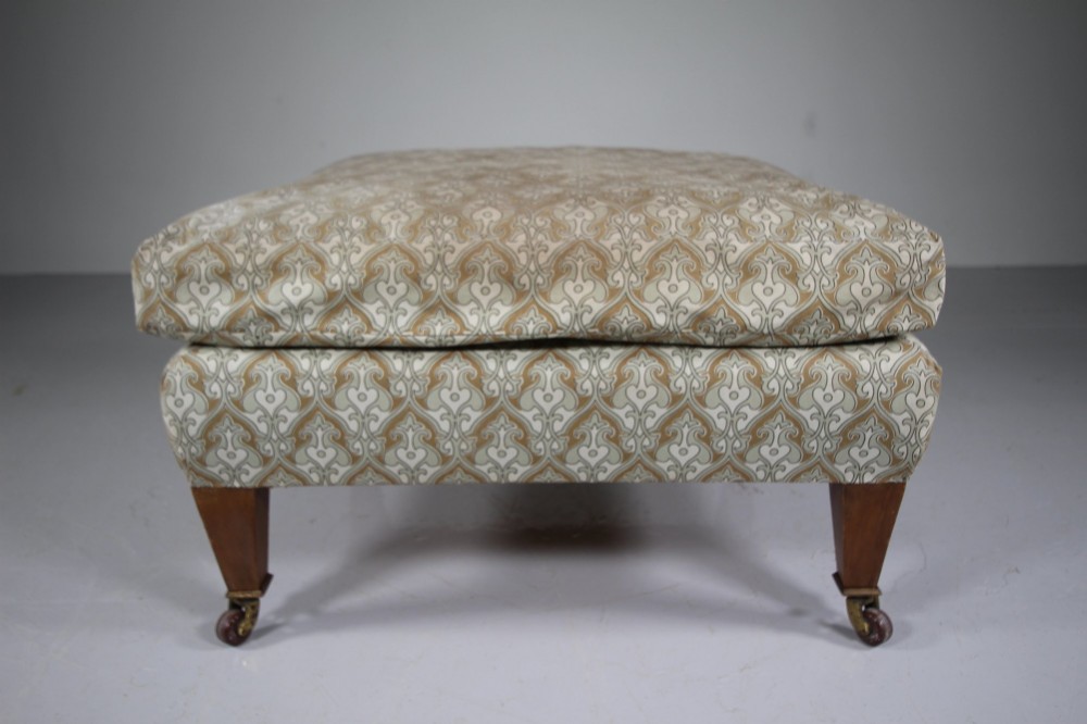 A large square antique footstool upholstered in a beige patterned fabric with a loose top cushion, supported by four tapered wooden legs on brass castors. View II