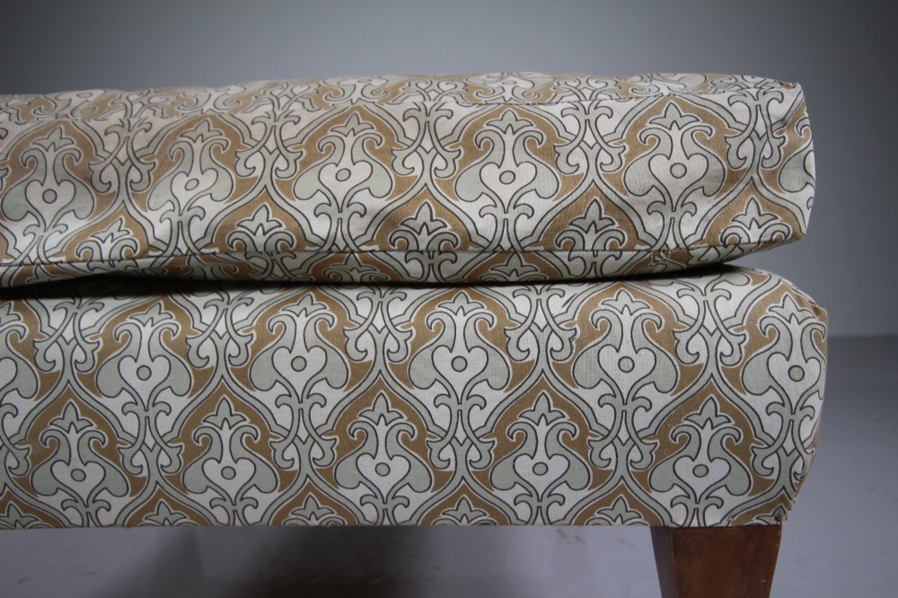 A large square antique footstool upholstered in a beige patterned fabric with a loose top cushion, supported by four tapered wooden legs on brass castors. View III