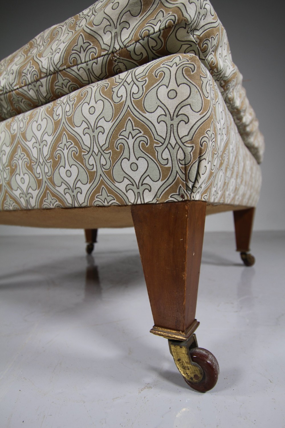 A large square antique footstool upholstered in a beige patterned fabric with a loose top cushion, supported by four tapered wooden legs on brass castors. View IV