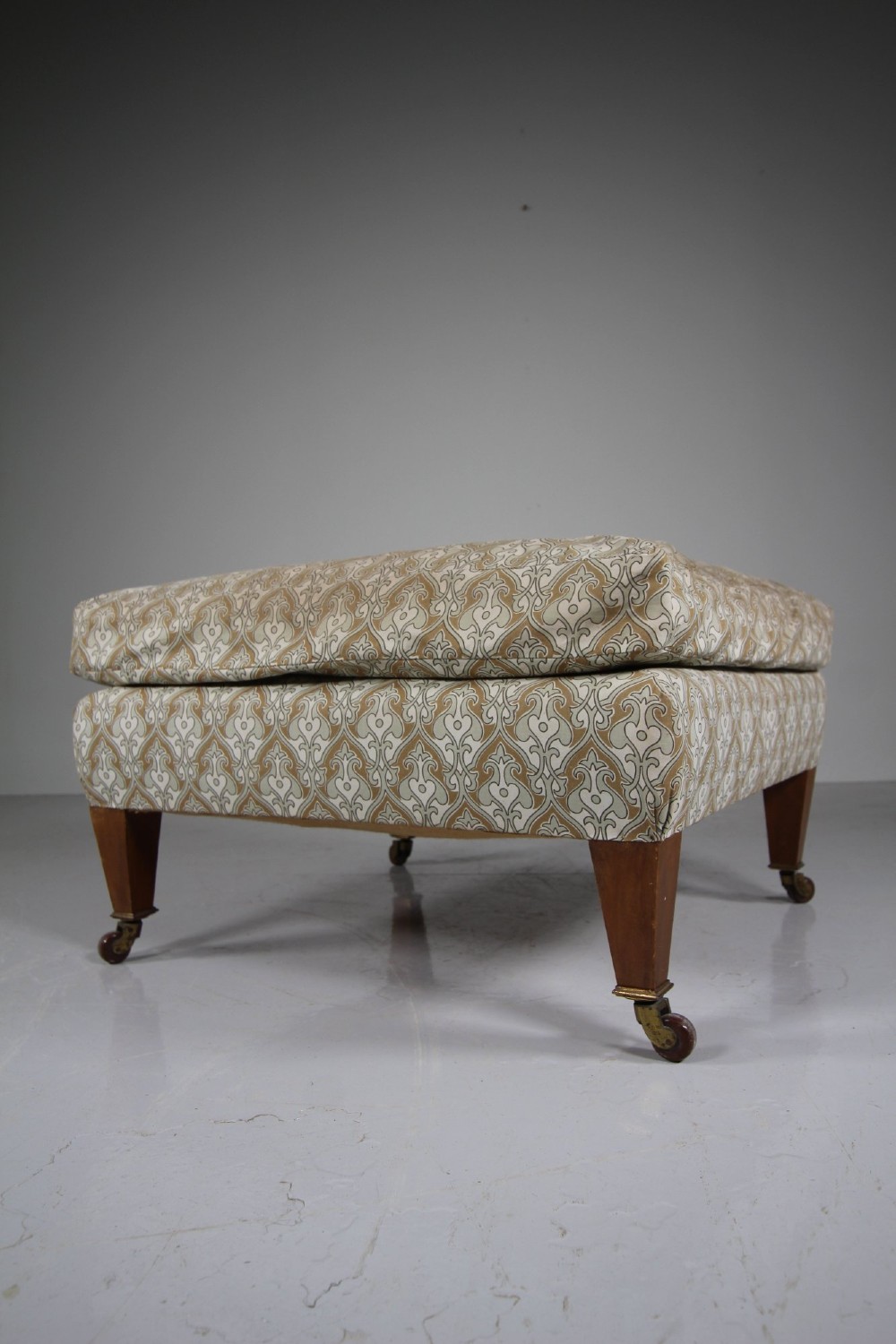 A large square antique footstool upholstered in a beige patterned fabric with a loose top cushion, supported by four tapered wooden legs on brass castors. View V