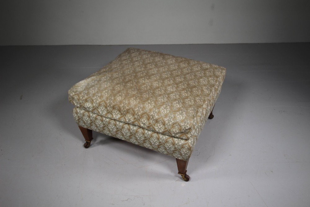 A large square antique footstool upholstered in a beige patterned fabric with a loose top cushion, supported by four tapered wooden legs on brass castors. View VI