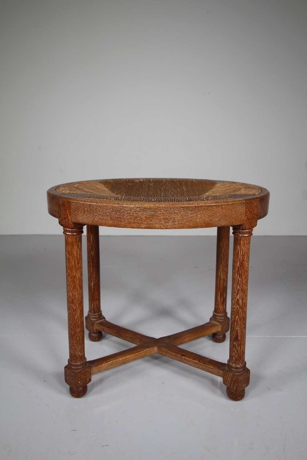 An antique oval oak stool featuring a woven natural rush seat, cylindrical turned legs, and a crossover X-frame stretcher at the base.