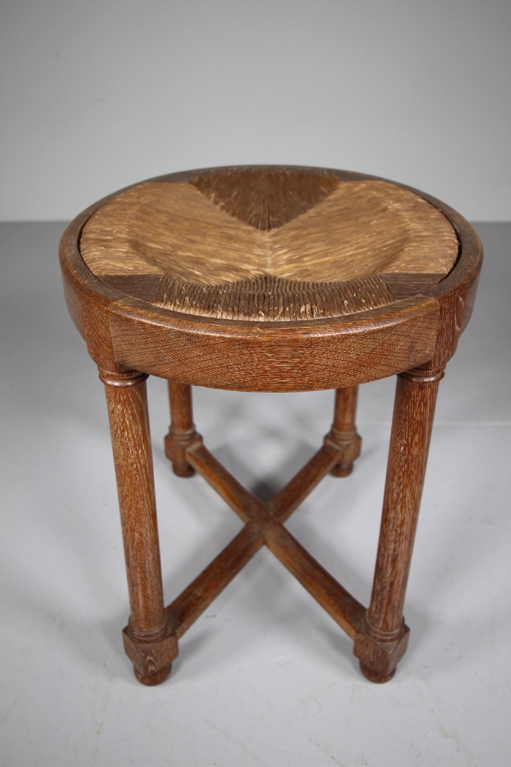 An antique oval oak stool featuring a woven natural rush seat, cylindrical turned legs, and a crossover X-frame stretcher at the base. View II