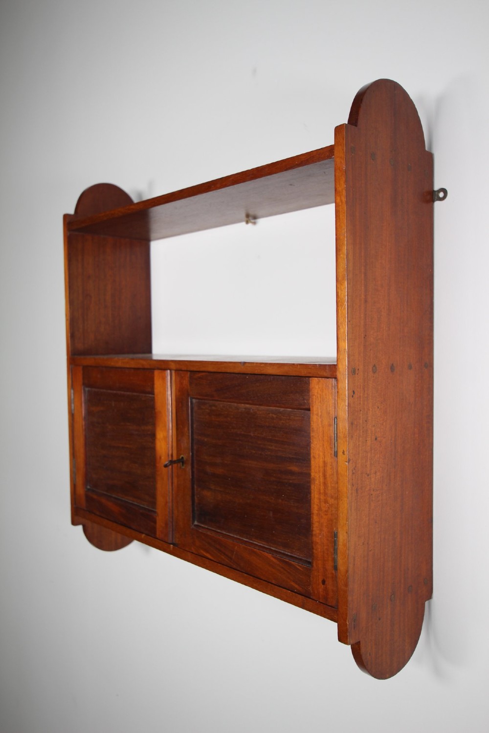 An Arts and Crafts walnut wall cabinet with an open top shelf above twin panelled cupboard doors and rounded side panels.