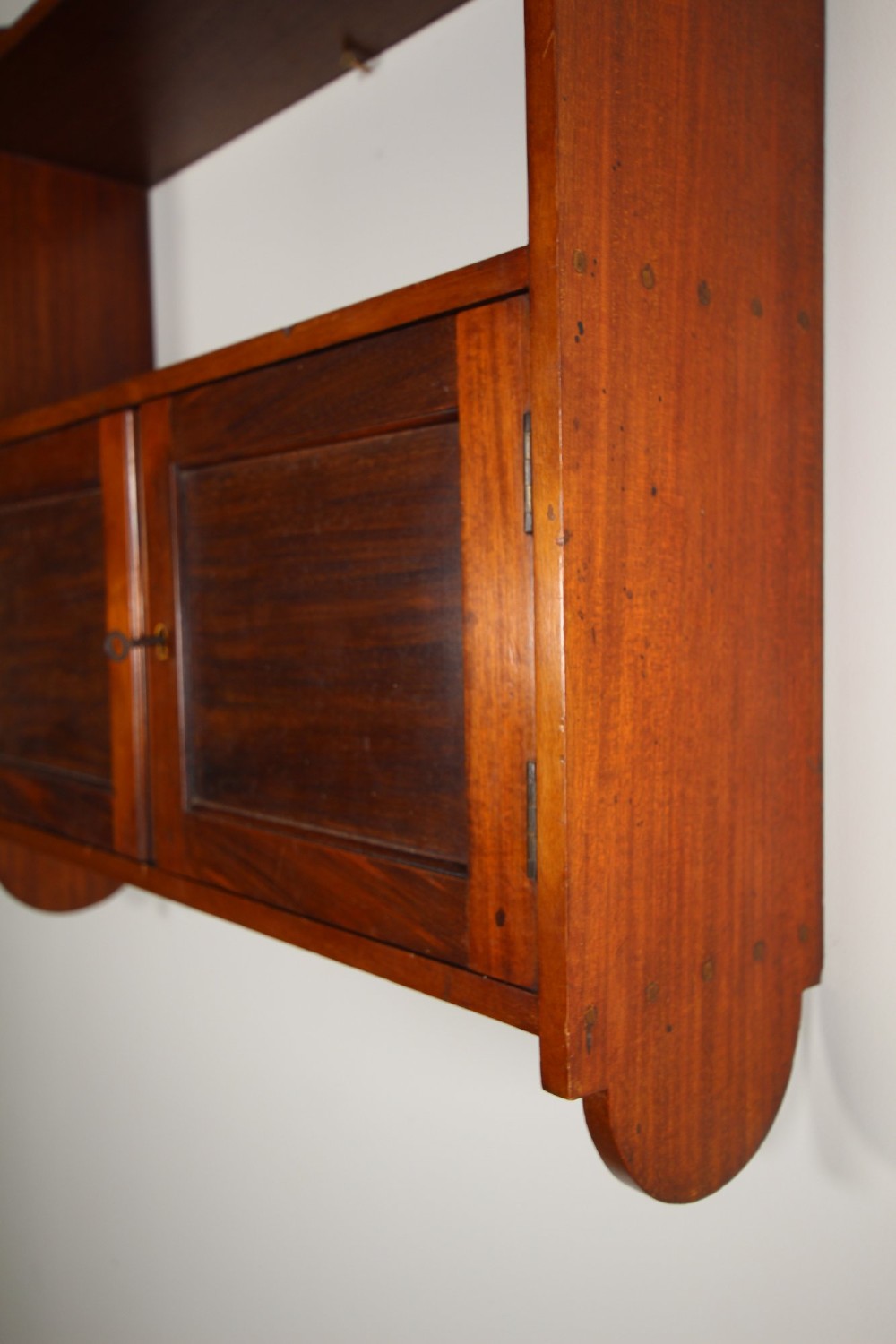 An Arts and Crafts walnut wall cabinet with an open top shelf above twin panelled cupboard doors and rounded side panels. View VII