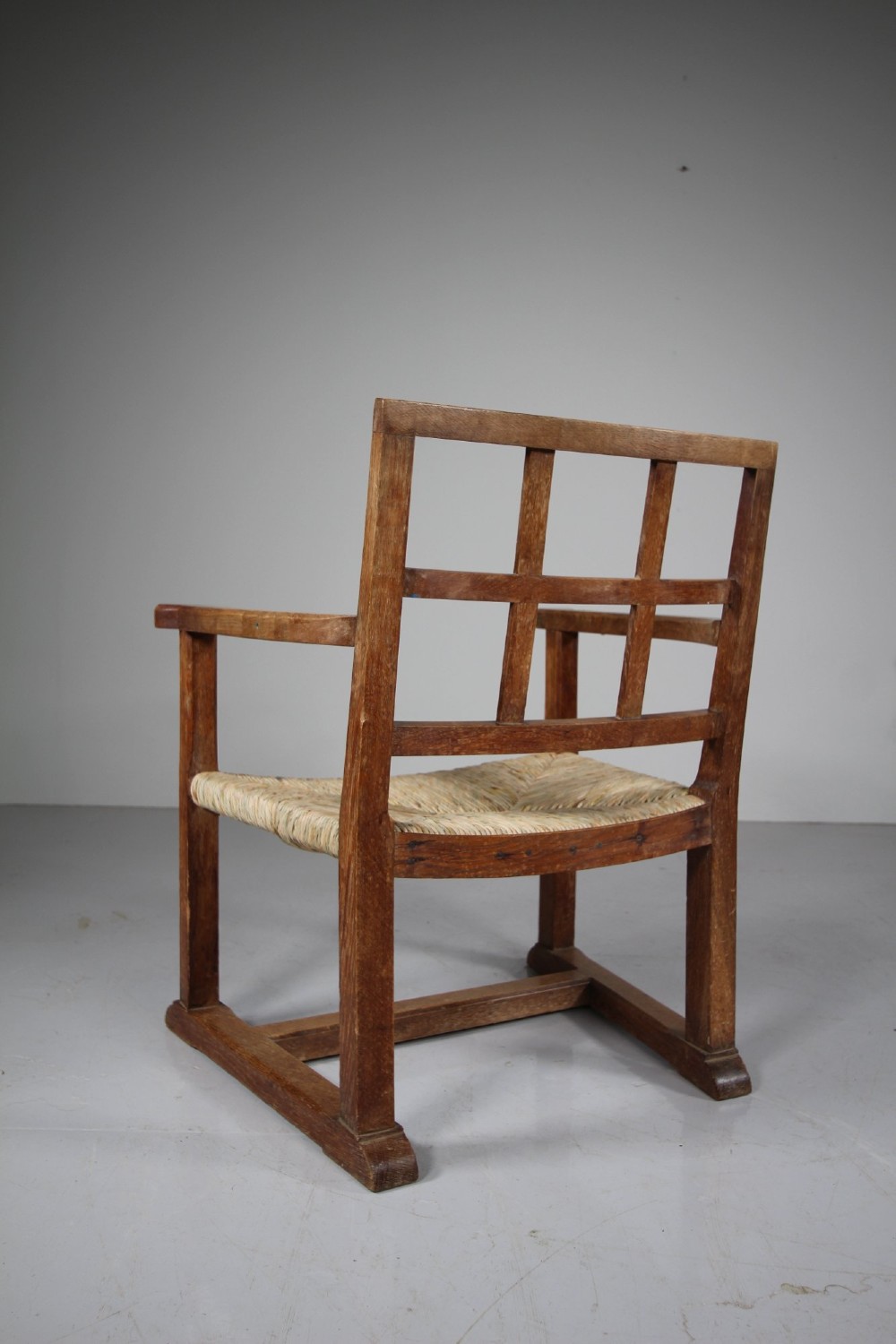 An Arts and Crafts oak fireside chair featuring a grid backrest, wide flat armrests, a woven rush seat, and a sledge base. View II