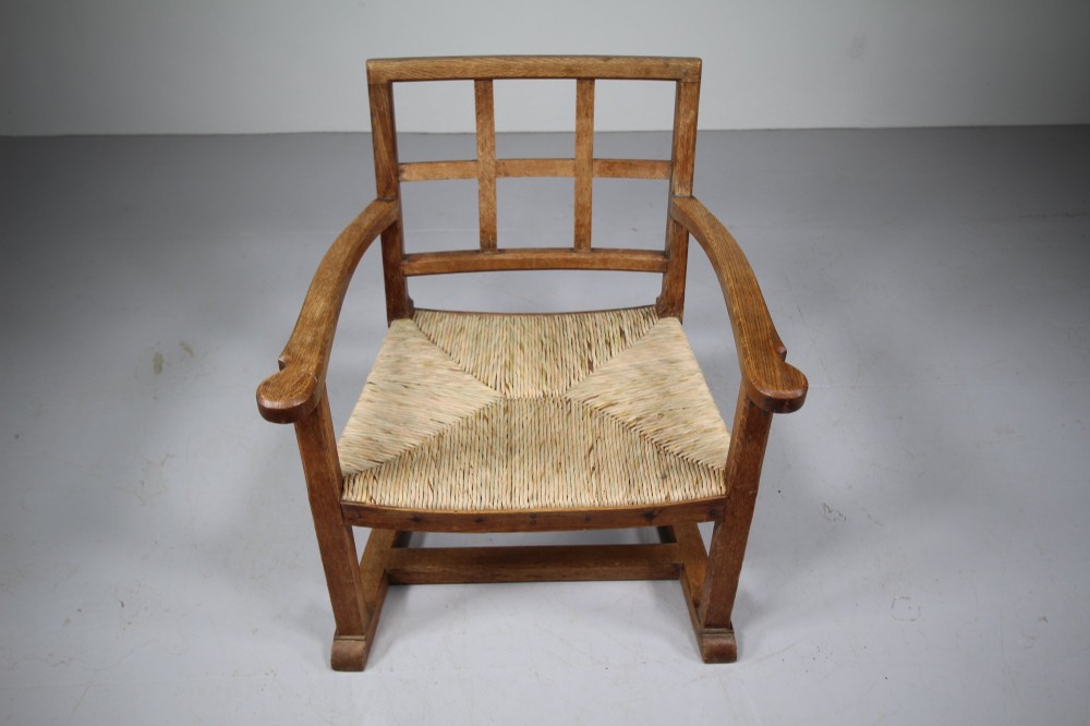 An Arts and Crafts oak fireside chair featuring a grid backrest, wide flat armrests, a woven rush seat, and a sledge base. View IV