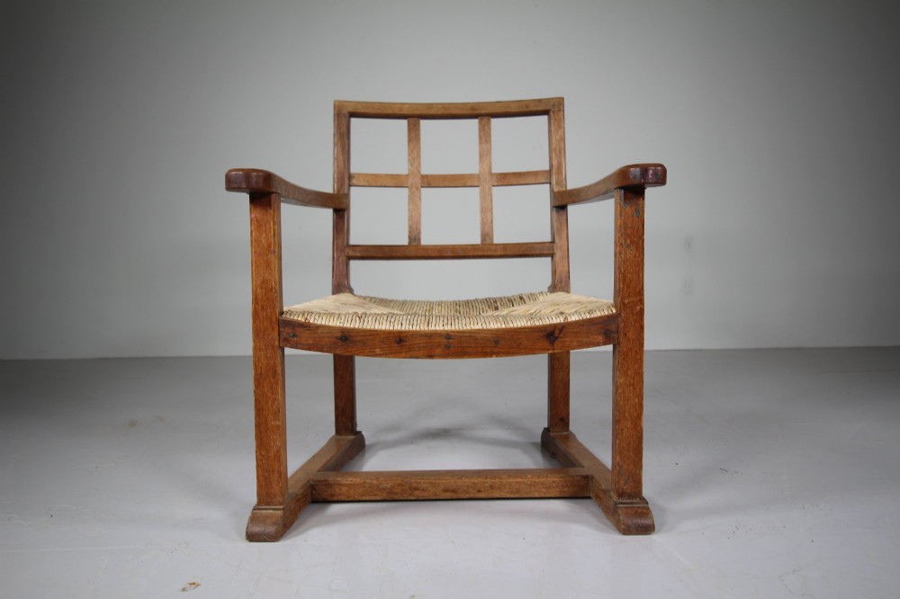 An Arts and Crafts oak fireside chair featuring a grid backrest, wide flat armrests, a woven rush seat, and a sledge base. View V