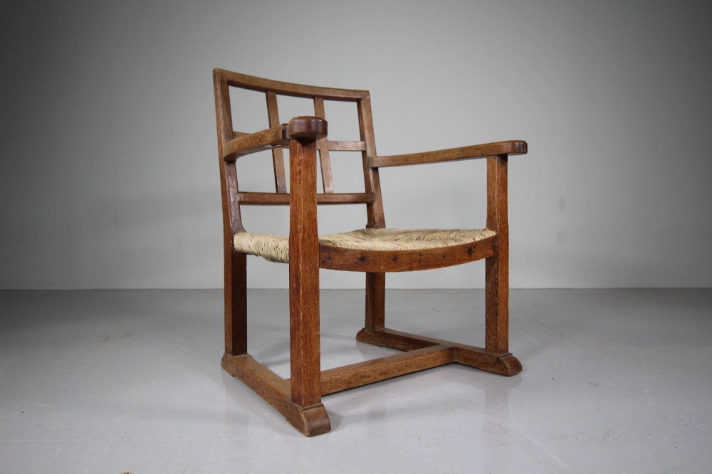 An Arts and Crafts oak fireside chair featuring a grid backrest, wide flat armrests, a woven rush seat, and a sledge base. View VI