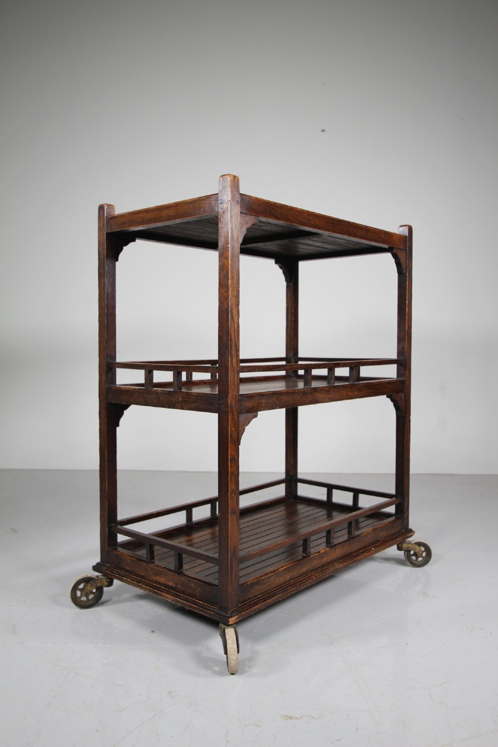 A dark oak three-tiered Edwardian trolley featuring slatted shelf galleries and castor wheels.