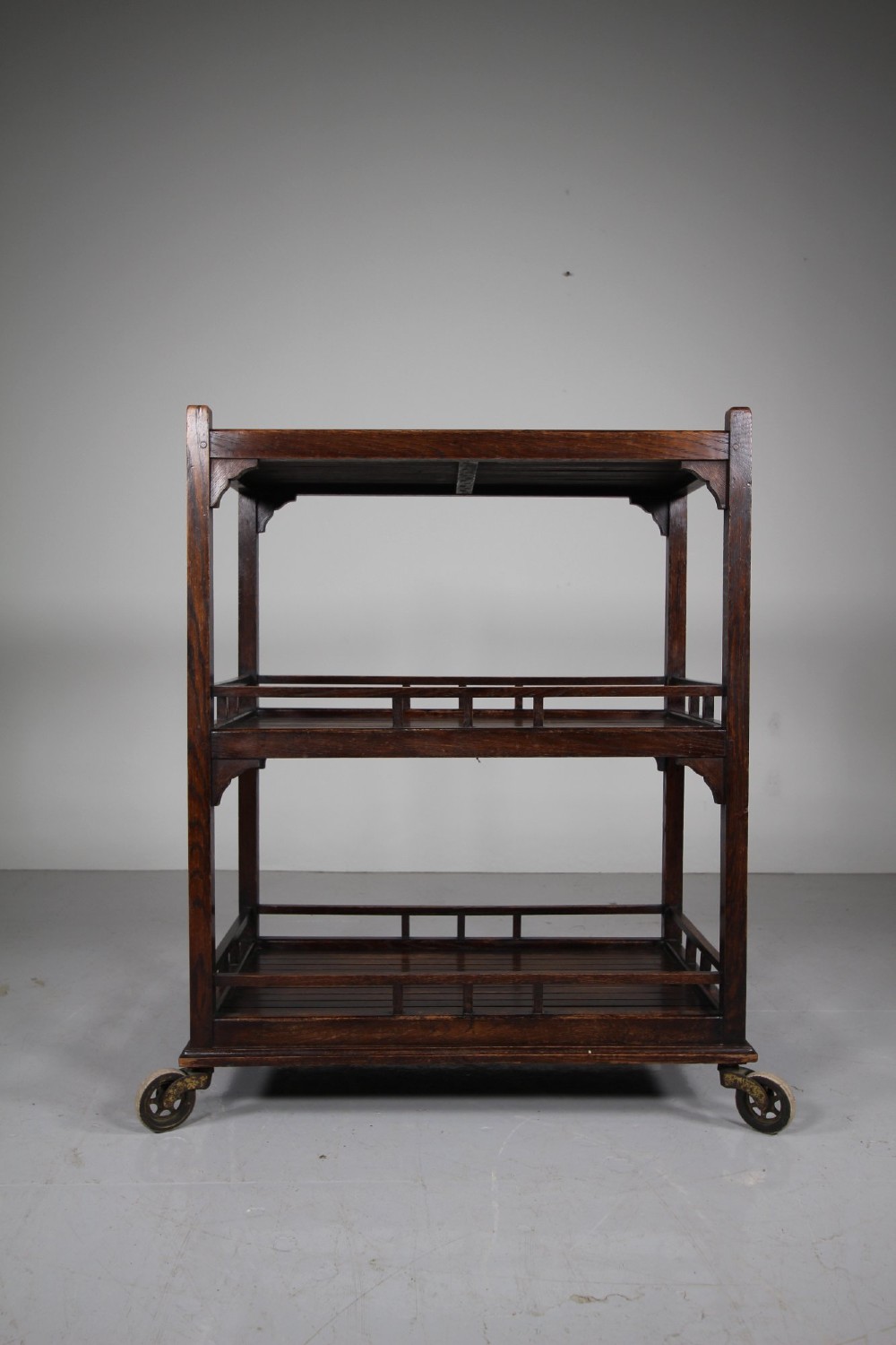 A dark oak three-tiered Edwardian trolley featuring slatted shelf galleries and castor wheels. View III