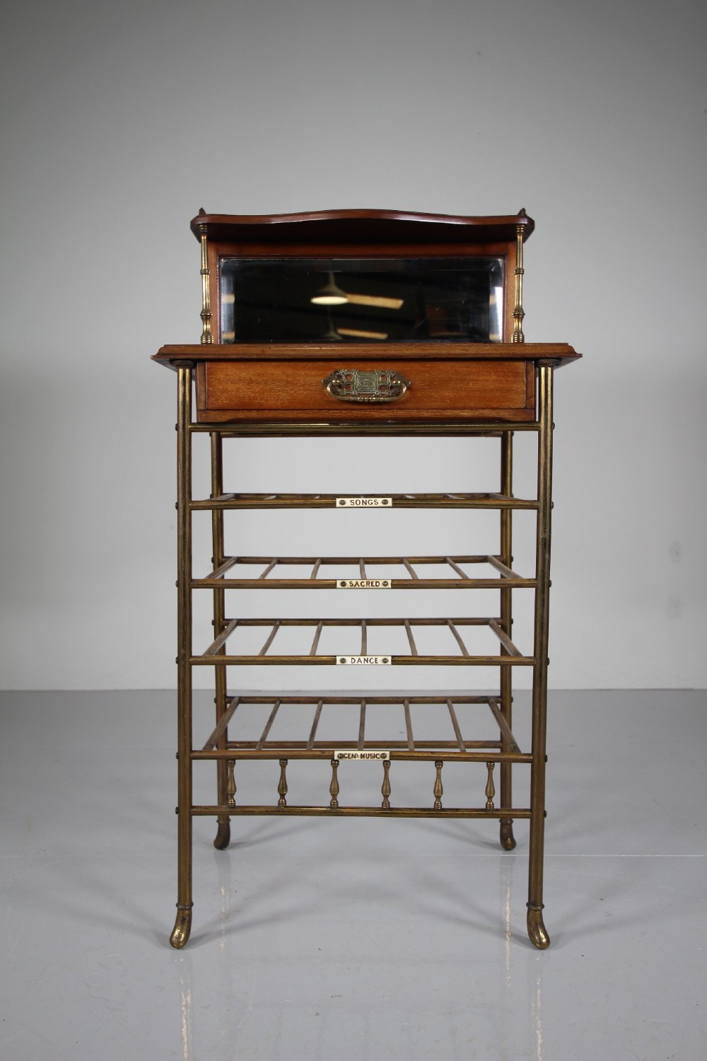 A 19th-century English Victorian brass and mahogany sheet music stand featuring open slatted shelves with ceramic labels, a top drawer, and a mirrored upper gallery.