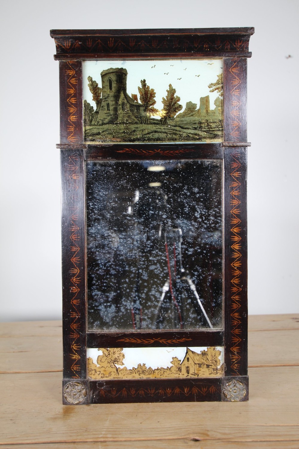 An early 19th-century rectangular wall mirror featuring a dark painted wooden frame with stencilled foliate borders and two reverse-painted glass panels displaying rural landscape scenes. View III