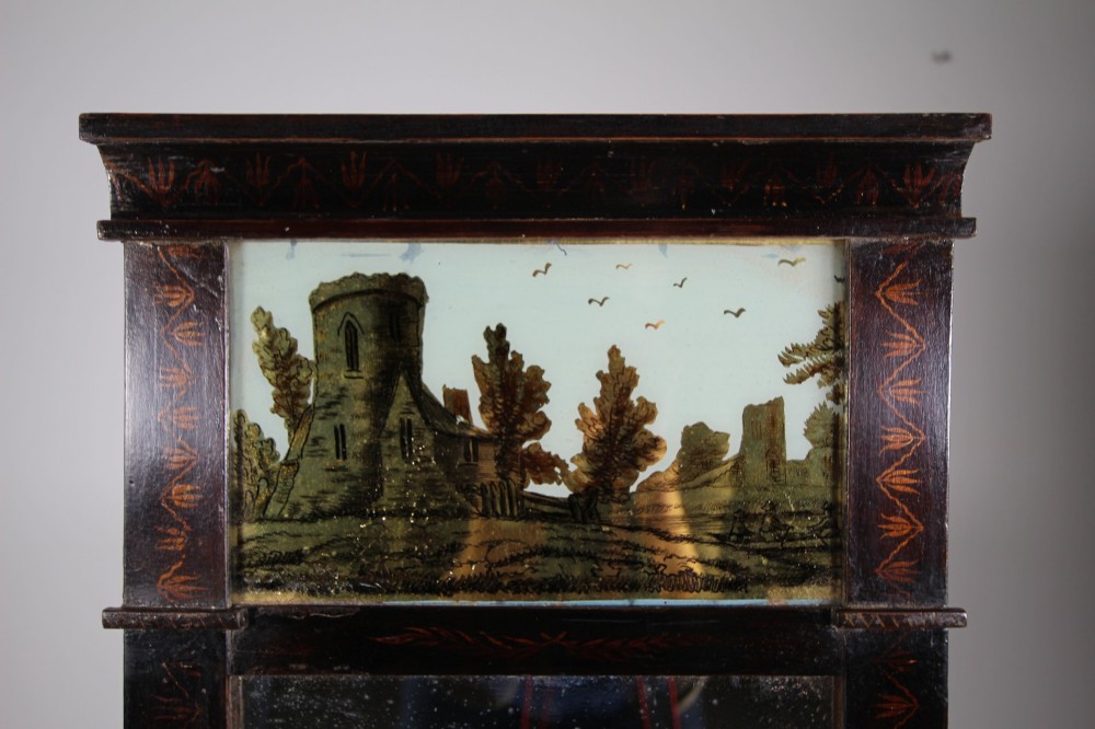 An early 19th-century rectangular wall mirror featuring a dark painted wooden frame with stencilled foliate borders and two reverse-painted glass panels displaying rural landscape scenes. View IV