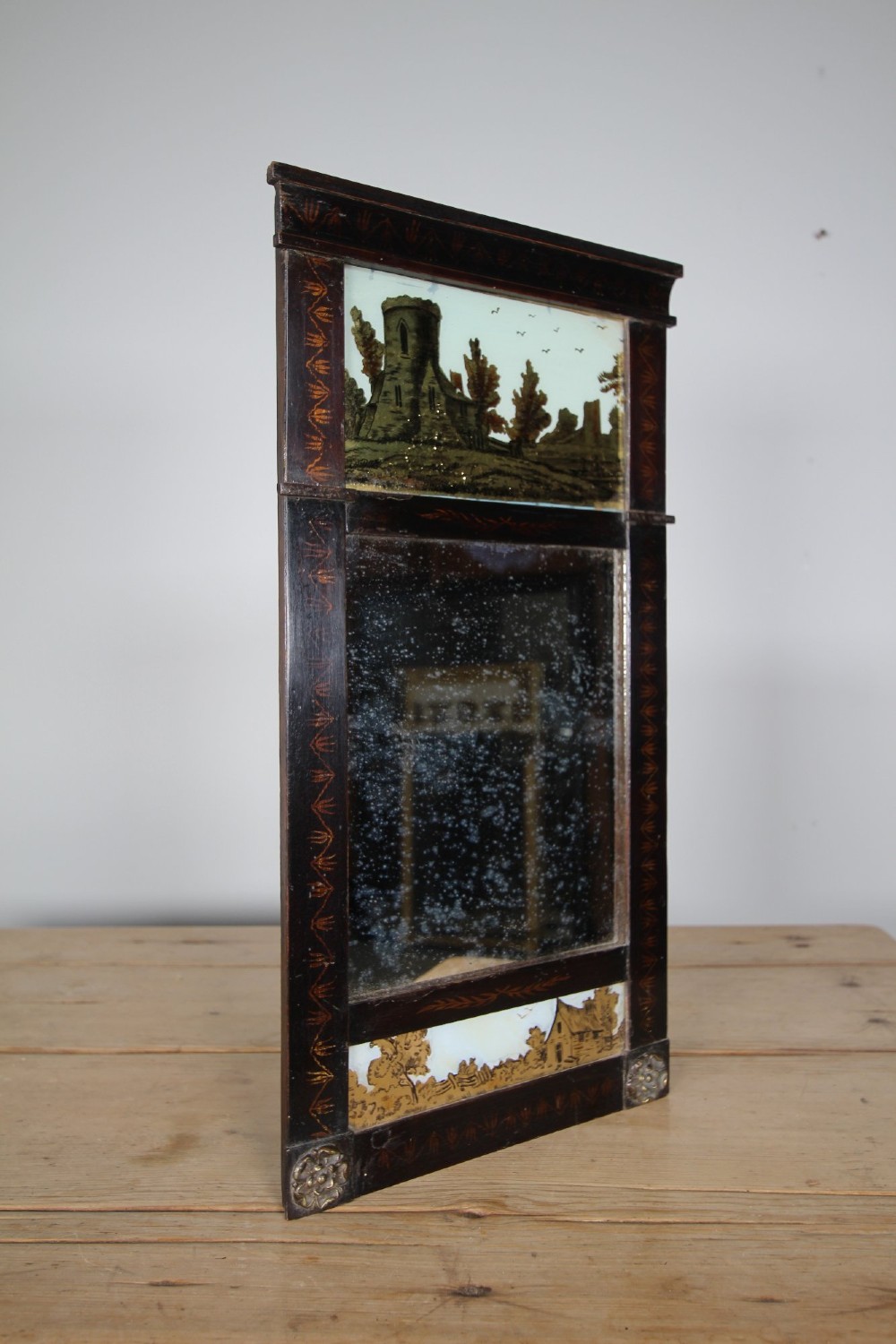 An early 19th-century rectangular wall mirror featuring a dark painted wooden frame with stencilled foliate borders and two reverse-painted glass panels displaying rural landscape scenes. View VIII