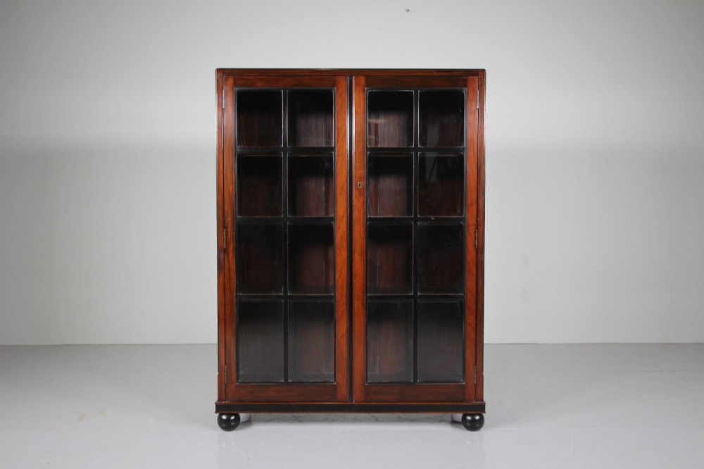 A 1920s Heals mahogany bookcase featuring two glazed doors with multi-pane glass and dark turned bun feet.