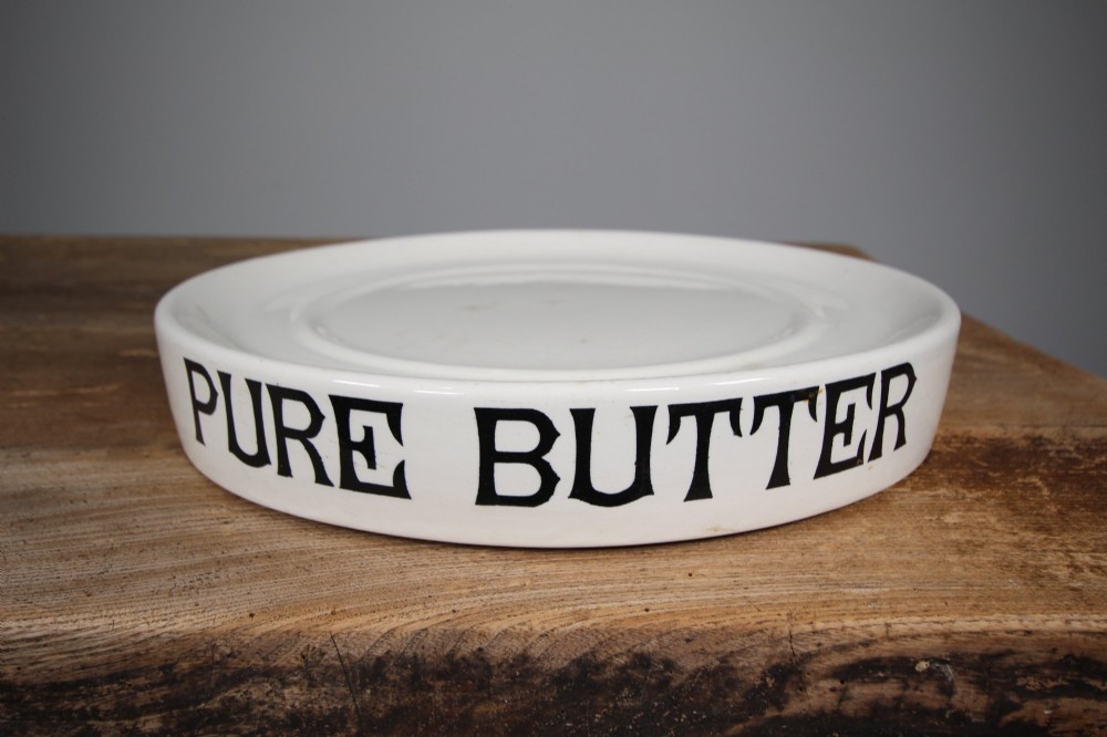 A circular creamware glazed ceramic butter slab featuring the words PURE BUTTER printed in black bold serif typography along the rim.