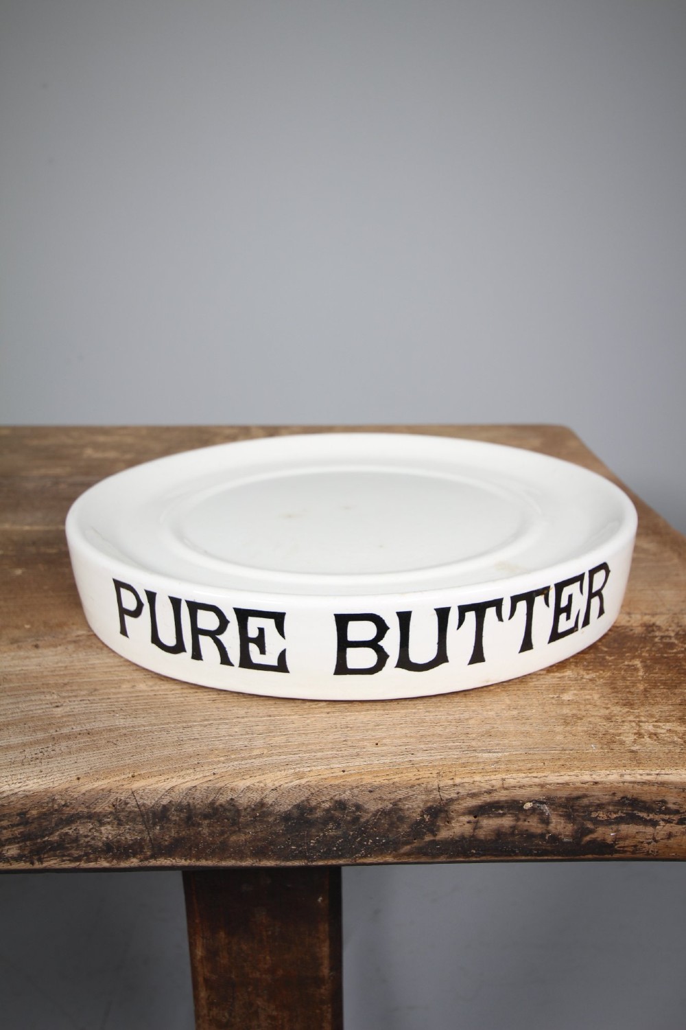 A circular creamware glazed ceramic butter slab featuring the words PURE BUTTER printed in black bold serif typography along the rim. View V