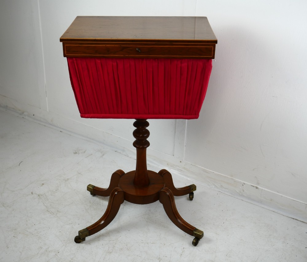 A Regency mahogany sewing table featuring a rectangular top with string inlay, a pleated red fabric work bin below, a turned pedestal support, and four splayed legs with brass castors.