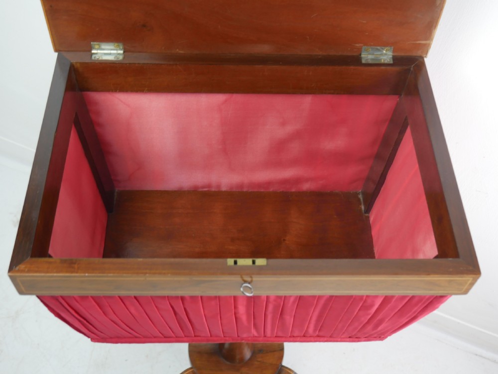 A Regency mahogany sewing table featuring a rectangular top with string inlay, a pleated red fabric work bin below, a turned pedestal support, and four splayed legs with brass castors. View II