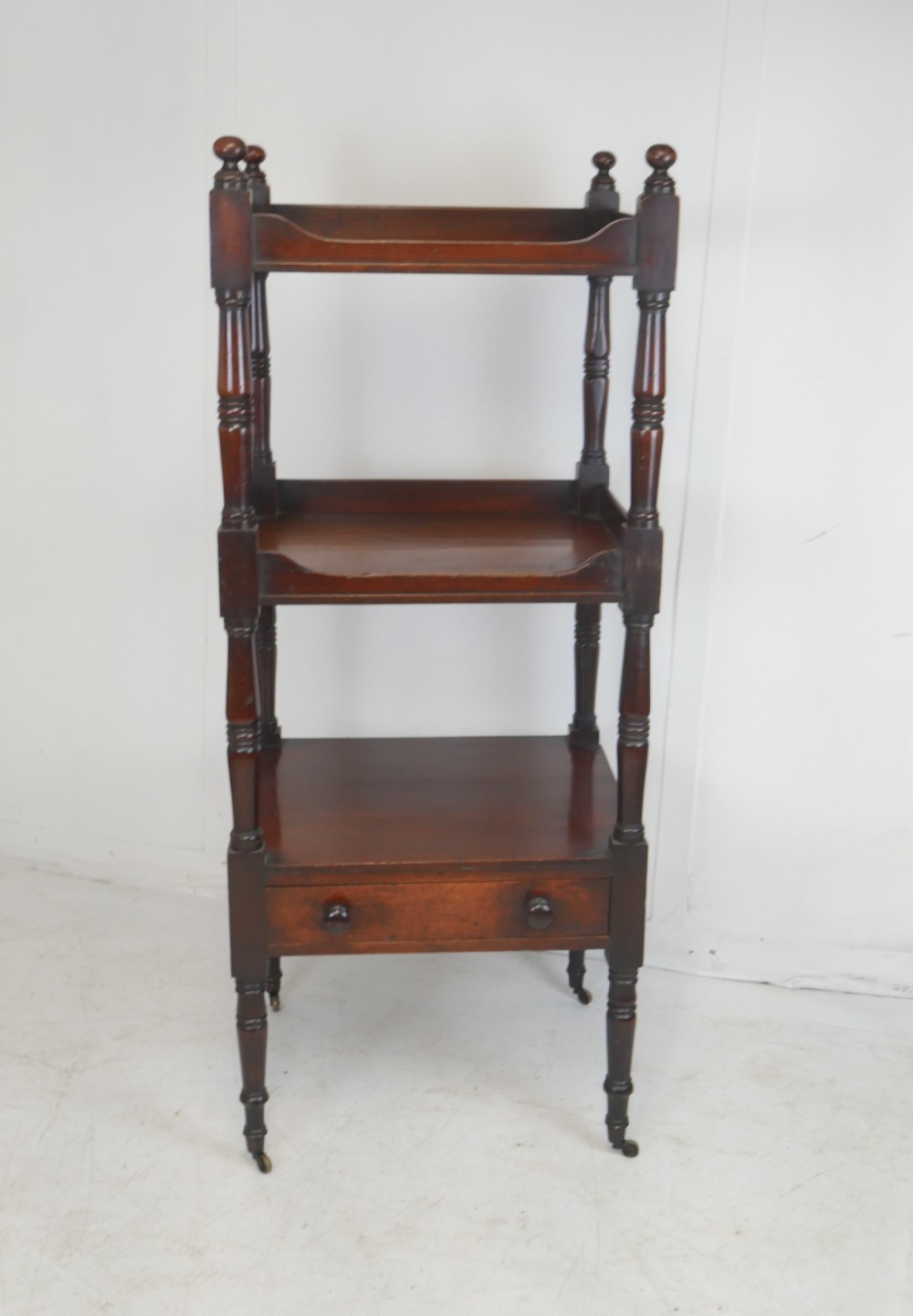 A three-tiered Regency mahogany whatnot stand featuring turned side columns, corner finials, a single base drawer with wooden knobs, and brass castors.